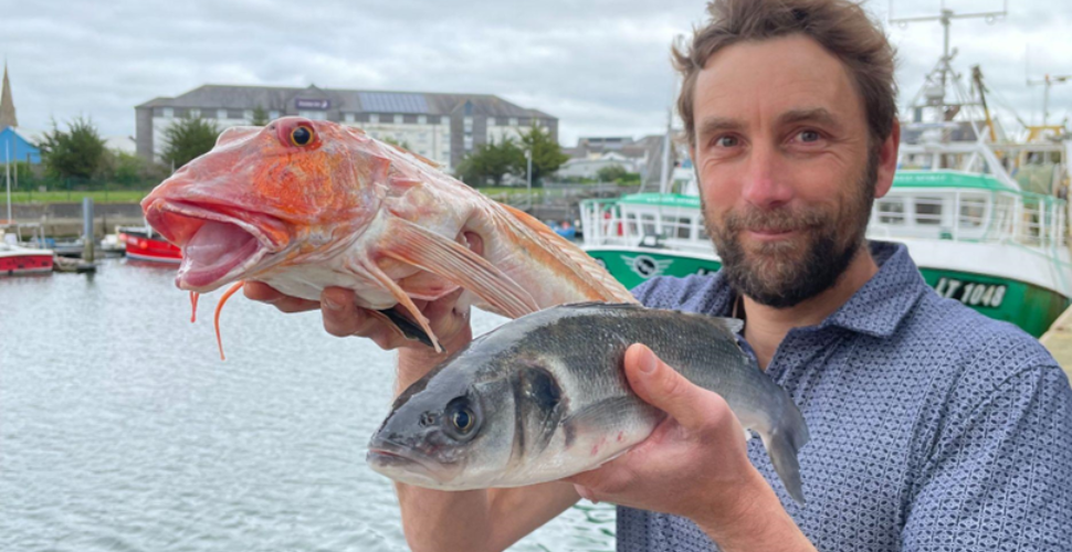 Man holding two fish up with a harbour in the background